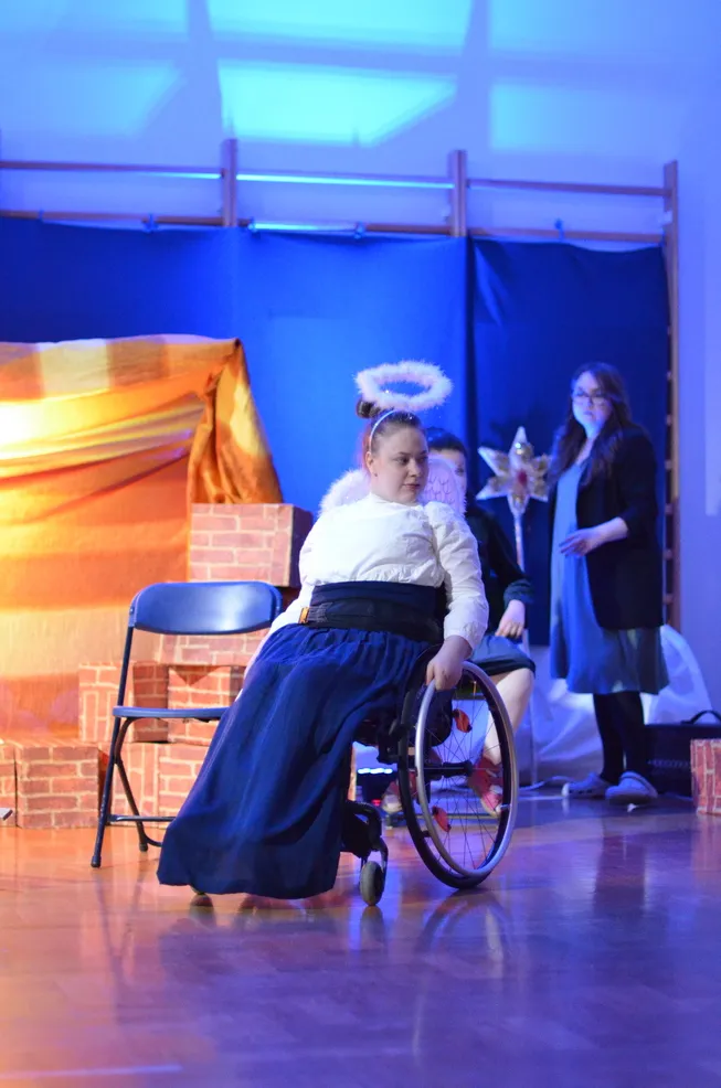 4. Uczennica jest ubrana w białą bluzkę, granatową, długą spodnicę, a na głowie ma aureolę. W tle widać nauczycielkę, szopkę i Gwiazdę Betlejemską