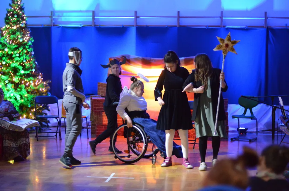 5. Uczniowie występujący w Jasełkach wędrują do Betlejem. Nauczycielka trzyma Gwiazdę Betlejemską. W tle widać szopkę i choinkę