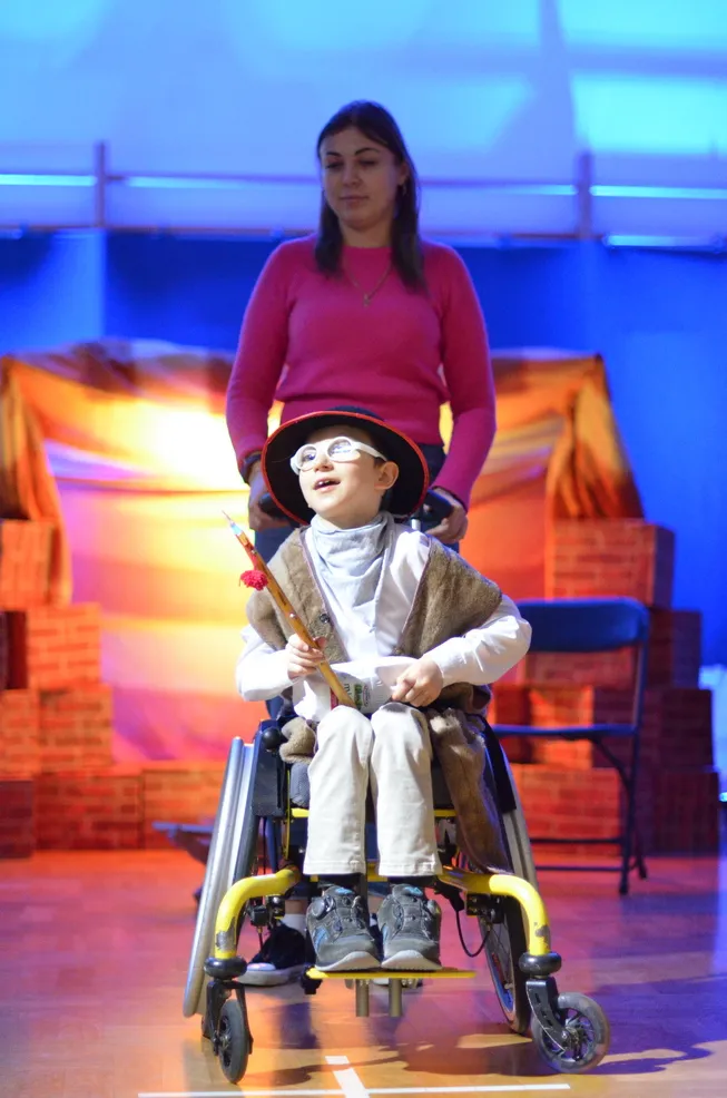 6. Uczeń przebrany za pastuszka ma na głowie kapelusz, a w ręku trzyma ciupagę. Za chłopcem, na tle szopki, stoi Pani Asystentka