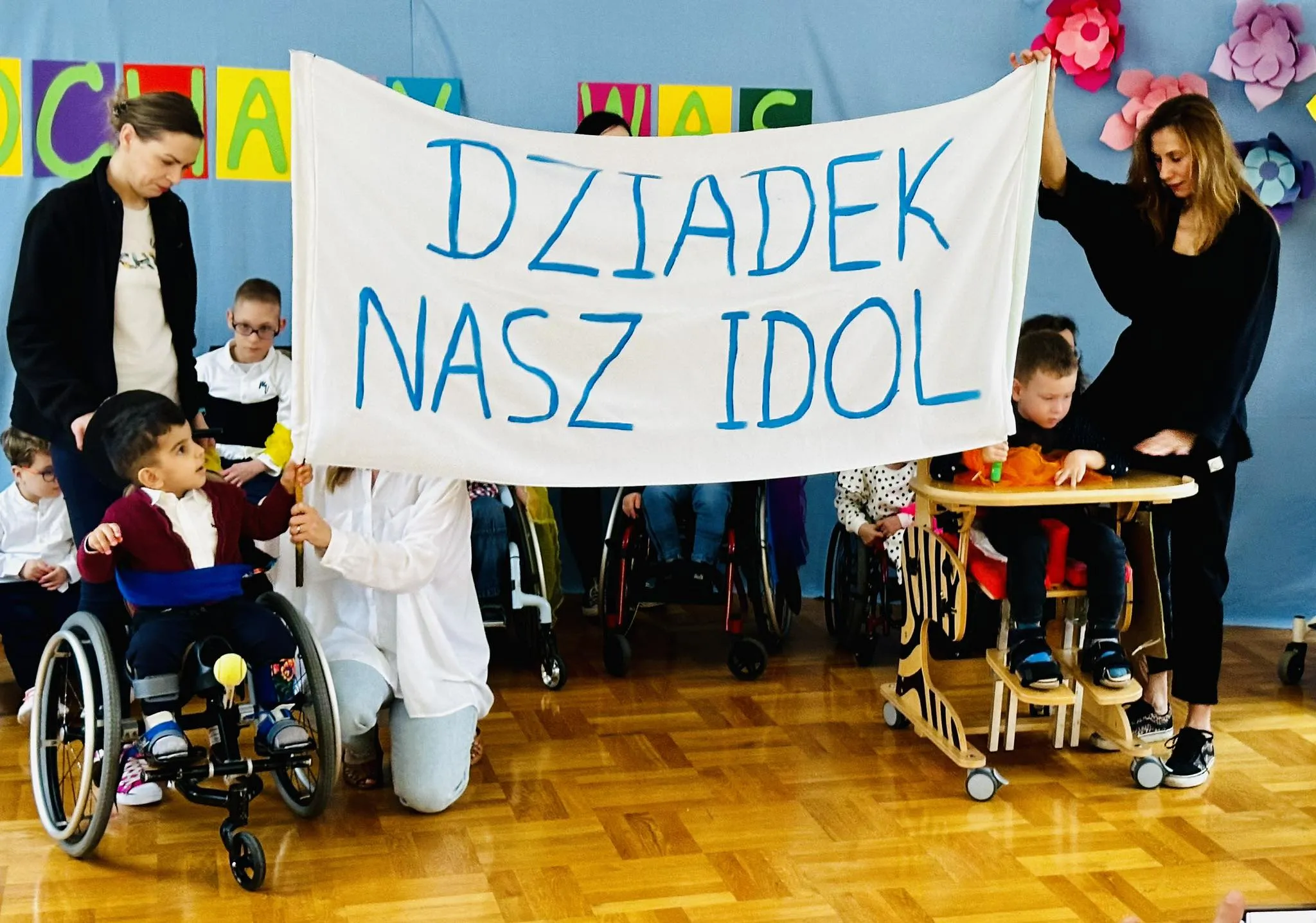 Dzieci z pomocą nauczycielek podczas przedstawienia trzymają transparent z napisem „DZIADEK NASZ IDOL”